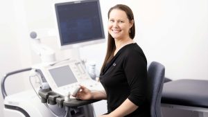 Female Sonographer facing towards you smiling sitting down at an ultrasound machine for X-ray Imaging Kawana